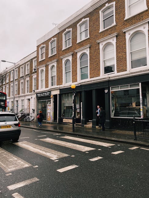 Street view of a narrow pavement in West Kensington featuring a row of commercial buildings with large glass windows and decorative black wrought iron balconies above. The shops have pastel green and 
