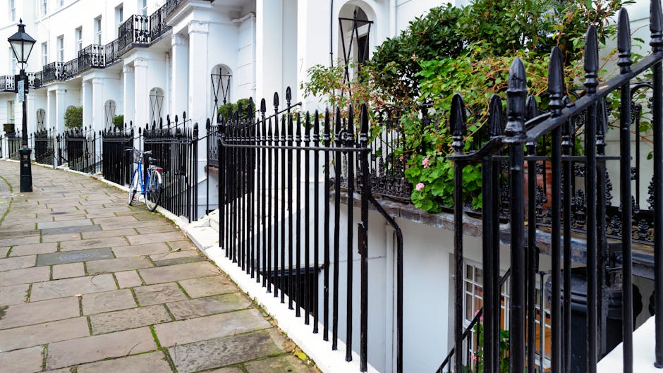 A narrow residential street with a white, multi-storey building featuring black wrought-iron balconies and window grilles. The pavement is made of rectangular stone slabs, some with visible moss or dirt. A black metal gate runs along the edge of the sidewalk, with some sections slightly open. A blue bicycle is parked and secured to the gate on the left side near a downward staircase leading to a basement entrance. Green foliage and pink flowers grow along the ledge adjacent to the building, adding contrast to the urban scene. The setting reflects a typical neighbourhood where house removals or moving services by Man and Van West Kensington may be conducted, with space for loading furniture and boxes from property into parked vehicles for home relocation.