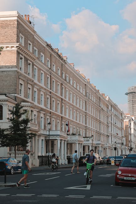 The image shows a row of historic terraced houses typical of West Kensington, featuring pale brick facades, decorative cornices, and traditional sash windows. The buildings are multi-story, with shops or vestibules on the ground floor, and are set along a wide street under a partly cloudy sky. Several pedestrians are walking along the pavement, while a cyclist and a motorist are navigating the road. In the foreground, parked and moving cars are visible, including a red vehicle on the right. The scene suggests an urban environment suitable for house removals or relocation services. Occasionally, Man and Van West Kensington might be called upon for house removal projects in this area, which often involves packing, loading, and transportation of household furniture and belongings through busy streets such as this, with careful attention to space management and logistics amidst urban traffic. The overall setting reflects a typical West Kensington neighbourhood scene, emphasizing the context of home relocation or furniture transport.