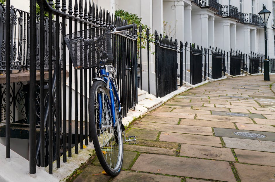 A blue bicycle with a front metal wire basket parked upright against a black wrought iron fence outside a white residential building with decorative columns and balconies in West Kensington. The sidewalk consists of large, uneven stone slabs, with some patches of moss or dirt between them. Several manhole covers are visible along the pavement. The scene is well-lit with natural daylight, casting minimal shadows, illustrating an outdoor setting typical for home relocation or furniture transport in urban environments. The presence of the bicycle leaning against the fence indicates possible use for moving or errands, while the close proximity to the building suggests access for loading boxes, furniture, or appliances during a house removal. The image supports content about common logistical challenges in removals from flats within West Kensington, as handled by companies like Man and Van West Kensington.