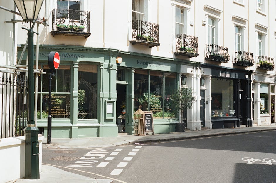 The image shows a row of historic terraced houses typical of West Kensington, featuring pale brick facades, decorative cornices, and traditional sash windows. The buildings are multi-story, with shops or vestibules on the ground floor, and are set along a wide street under a partly cloudy sky. Several pedestrians are walking along the pavement, while a cyclist and a motorist are navigating the road. In the foreground, parked and moving cars are visible, including a red vehicle on the right. The scene suggests an urban environment suitable for house removals or relocation services. Occasionally, Man and Van West Kensington might be called upon for house removal projects in this area, which often involves packing, loading, and transportation of household furniture and belongings through busy streets such as this, with careful attention to space management and logistics amidst urban traffic. The overall setting reflects a typical West Kensington neighbourhood scene, emphasizing the context of home relocation or furniture transport.