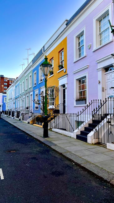 A row of colorful terraced houses along a residential street in West Kensington, with each property painted in pastel shades of blue, purple, yellow, and white, featuring large sash windows, decorativ