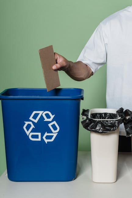 A man standing on a dirt area next to a large pile of assorted household waste and scrap metal, including plastic, wood, and electronic components, organized behind a brown metal fence with white lett
