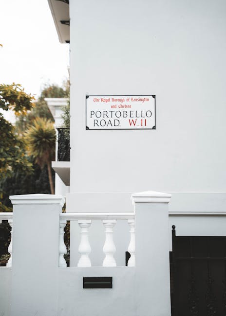 A white residential building with a small front terrace featuring a white balustrade and decorative columns, positioned on a pavement next to a street sign that reads 'PORTOBELLO ROAD W.11' and indicates the Royal Borough of Kensington and Chelsea. In the background, trees with green and autumnal foliage are visible, suggesting a suburban or urban area. The building's exterior includes a neatly painted white wall, partially visible windows, and a black gate. This scene is related to house removals and relocation services, illustrating a typical residential address where moving activities may take place, and a man and van company like Man and Van West Kensington may be required for furniture transport and packing during a home relocation process.
