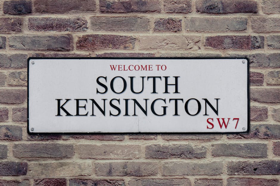 A rectangular white metal street sign mounted on a brick wall, displaying the text 'Welcome to South Kensington' in black uppercase letters, with 'SW7' in red at the bottom right corner; the sign is positioned at eye level on the brick exterior of a building, which features light brown and reddish bricks with visible mortar joints. The lighting is natural, illuminating the sign and the textured brick surface, suggesting the image was taken during daylight hours, in an urban area associated with house removals or relocation services such as those offered by Man and Van West Kensington.