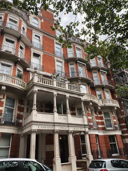 The exterior of a multi-storey residential building with red brick walls, white decorative stonework, and black wrought-iron balconies. The building features a prominent entrance with a classical-style portico supported by white columns, engraved with the address ‘13-36 CAMPDEN HILL COURT’. Surrounding the property, there are leafy trees with green foliage partly obscuring the upper floors. In the foreground, part of a street with parked cars is visible beneath the trees. This scene depicts the front view of a typical townhouse in West Kensington, relevant to house removals and home relocation services like those offered by Man and Van West Kensington, providing context for logistics involved in furniture transport and packing during moving projects.