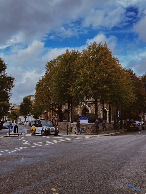 A street view showing a historic brick building with an arched entrance and a sign indicating 'Ladson Temple' on the front, surrounded by large trees with green and brown foliage. Several vehicles, including a small silver car and a black van, are parked along the curb, with some people walking on the pavement near the entrance. The scene captures a typical urban setting with overhead blue sky and clouds, and the surrounding area features a mixture of trees, street furniture, and traffic signage. This outdoor image provides context for house removals or home relocation services, highlighting the environment where [COMPANY_NAME] may operate for furniture transport and packing and moving activities near Olympia London in West Kensington.