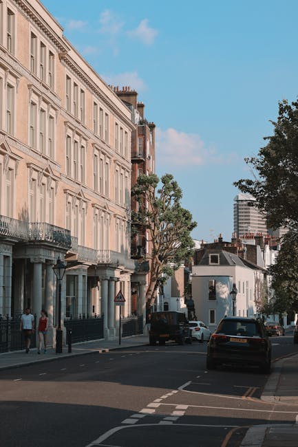 This image depicts a residential street in West Kensington, characterized by a row of cream-coloured, multi-storey Georgian-style houses with detailed cornices and balconies. The street scene is set during daylight, with a clear blue sky and a few scattered clouds. Several tall, leafy trees line the pavement, providing shade and greenery. Parked cars, including a dark vehicle and a white van, are positioned along the curbside, with some vehicles partially obscured by the trees. Pedestrians are seen walking along the sidewalk, passing by the houses and entering or exiting buildings. The street appears calm and orderly, suitable for home relocation or furniture transport activities as facilitated by local removals services. Occasionally, [COMPANY_NAME] may use such streets for loading and unloading furniture during house removals, ensuring careful handling of household items and efficient movement logistics in an urban environment typical of West Kensington.