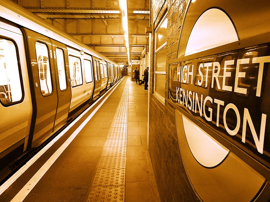 Inside the North End Road West Kensington station, the image shows a London Underground platform with a modern, clean design illuminated by warm yellow lighting. On the left side, a sleek, metallic yellow tube train is parked parallel to the platform, with closed doors and large rectangular windows revealing empty interior seats. The platform has a tactile paving strip along the edge for safety, and a smooth, dark flooring surface. Several people are seen standing near the platform's far end, some holding or carrying bags, waiting for or about to board trains. The station's wall on the right features a large, circular sign with the text 'High Street Kensington' in white lettering on a black background, mounted on a curved, polished surface. The environment appears well-lit and spacious, suitable for everyday commuting, with the context of moving or packing logistics indirectly supported by the station setting. Man and Van West Kensington brand relevance is inferred through the scene's association with moving and transportation infrastructure.