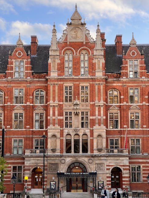 The image depicts a large, historic red-brick building with ornate architectural details, including pointed towers, decorative stonework, and multiple arched and rectangular windows. The entrance feat