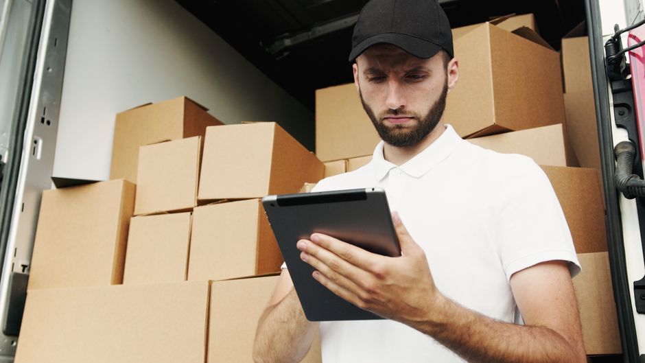 A person in light-colored clothing is seen inside a residential property, carefully lifting and stacking medium-sized cardboard boxes in preparation for a home relocation. The boxes are plain, brown, 