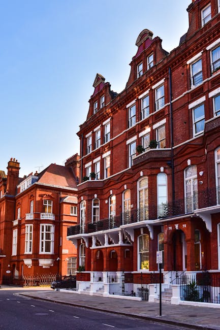 A row of red-brick Victorian residential buildings on Lillie Road in West Kensington, with large windows, decorative white trim, and black wrought-iron balconies. The ground level features staircases with white railings leading up to entrance doors, and the street in front has parked cars and a signpost. The image is taken during daylight with clear blue sky overhead. This setting depicts a typical location for house removals and furniture transport, reflecting the environment where Man and Van West Kensington performs home relocation and moving services, including loading and packing processes in an urban residential area.